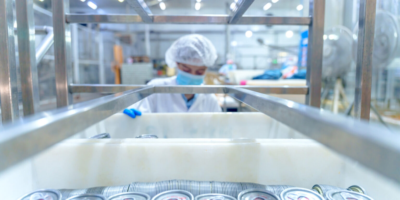 A worker in a food processing plant inspects metal cans. The scene shows shelves filled with stacked cans. The worker wears a mask and hairnet while focusing on the task during a busy day.